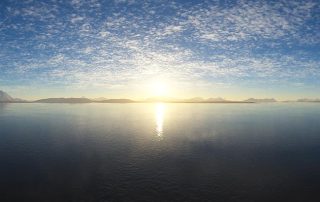 A panoramic photo of the sun rising across water with a strip of land in the distance and clouds in the sky.
